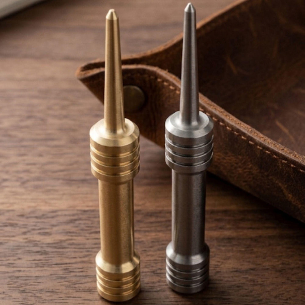 Brass and stainless steel barbell single prong divot tools standing on a wood table beside a leather tray